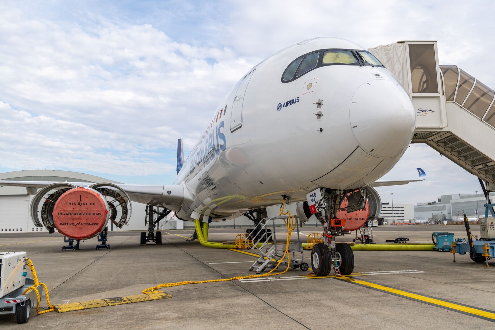 Nez d’un A350 Airbus blanc sur le tarmac, moteurs bâchés orange, passerelle Solari à droite
