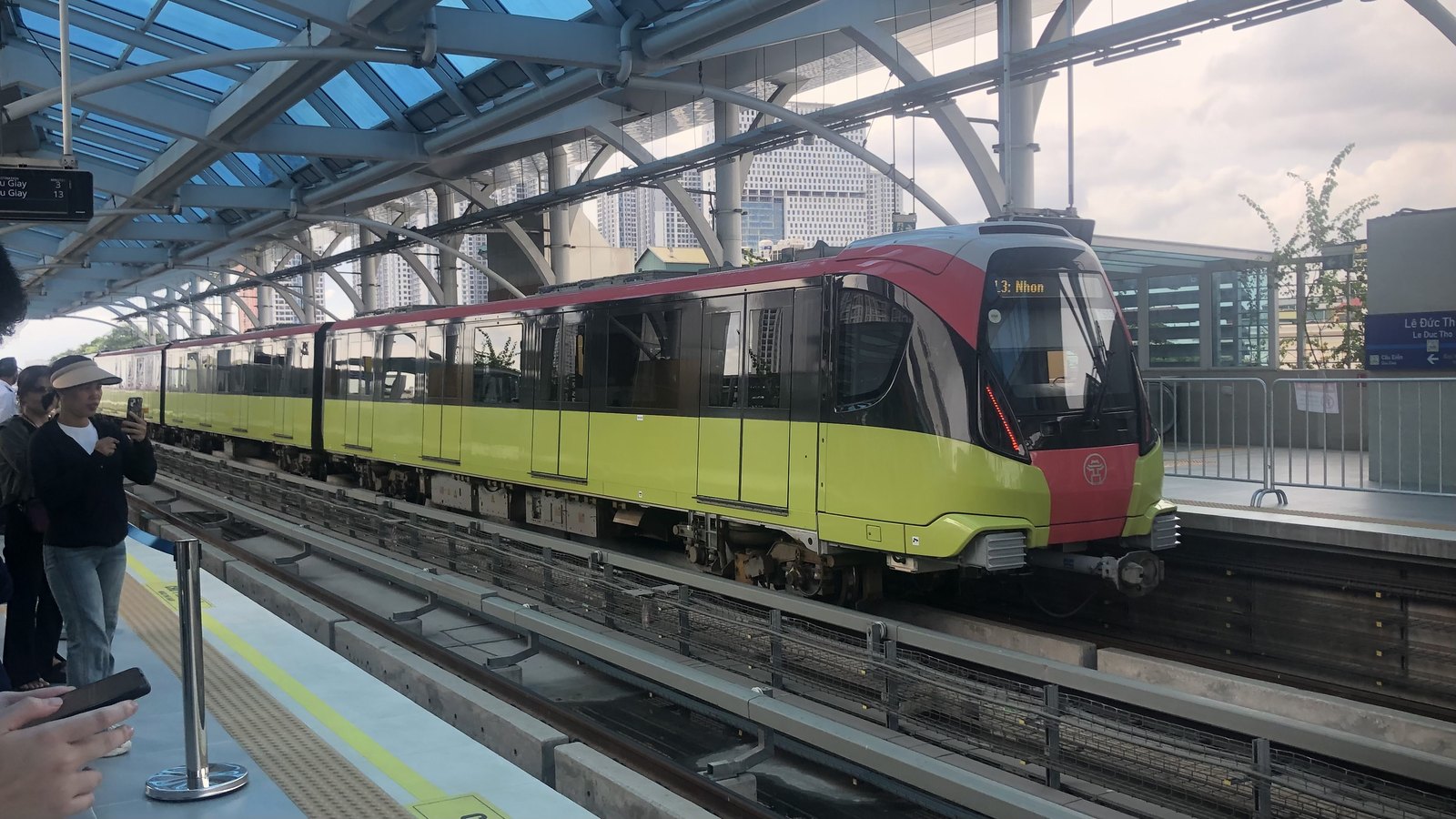 Une rame Metropolis jaune et rouge à quai dans une station aérienne de Hanoï, des voyageurs la photographient au premier plan