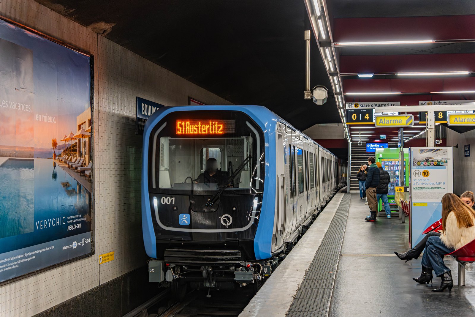 Une rame MF19 blanche et bleue, numéro 001, à quai dans une station du métro parisien, des voyageurs assis sur le quai