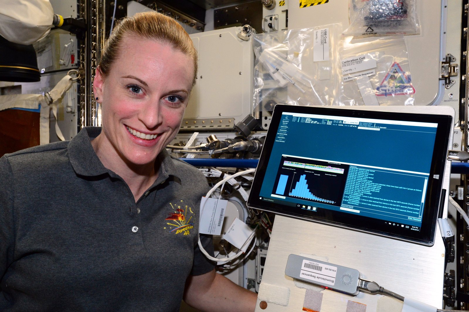 L’astronaute Kate Rubins, souriante, à côté d’un ordinateur relié à un petit séquenceur d’ADN à bord de la Station spatiale internationale
