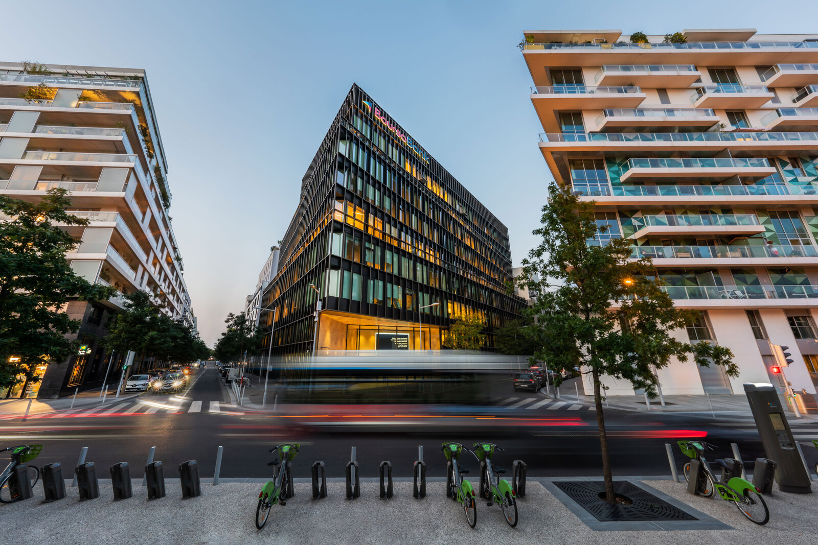 Immeuble de bureaux aux façades vitrées colorées au crépuscule, entouré de résidences à balcons et d’une rangée de vélos