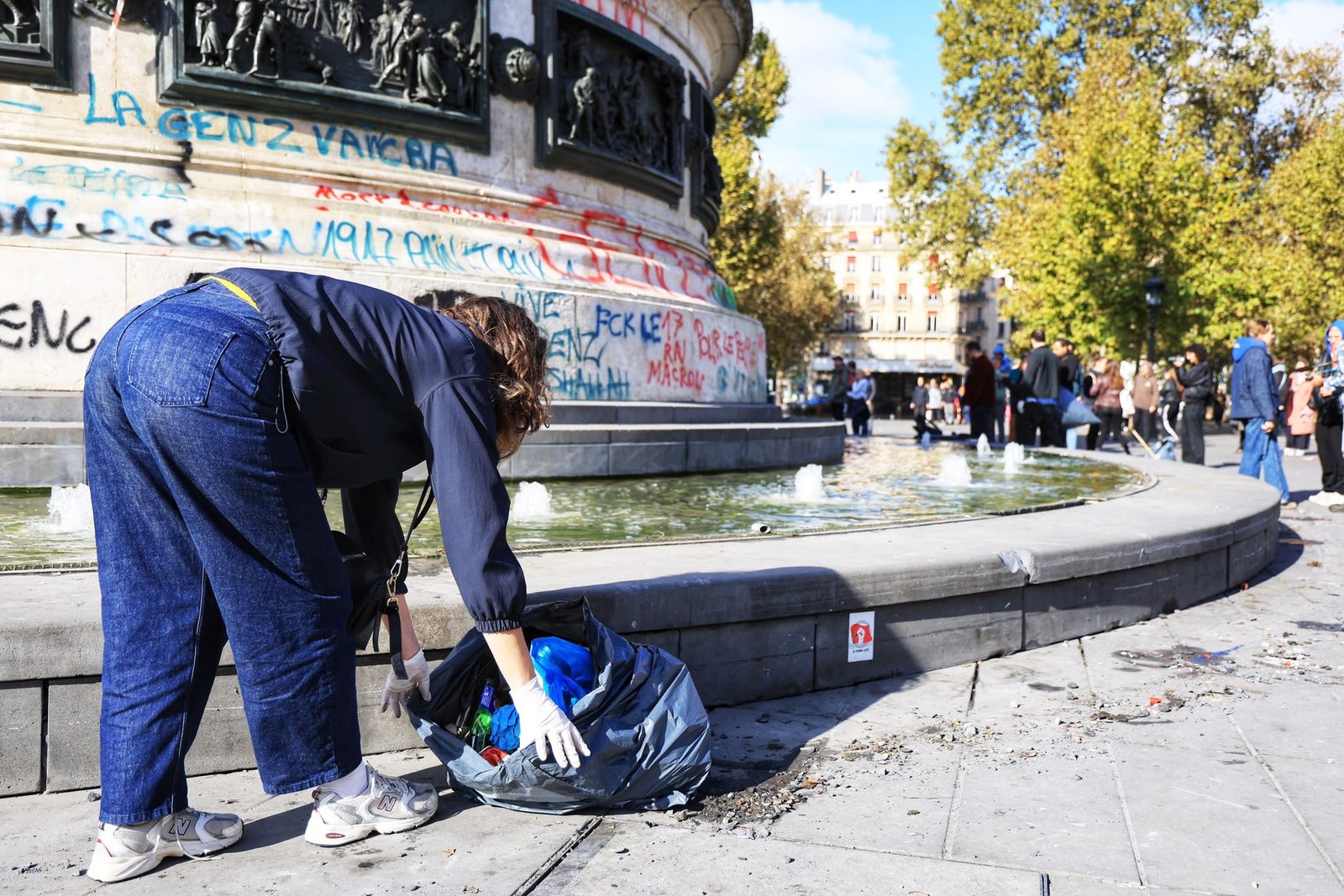 Un homme en veste bordeaux et gants blancs ramasse des débris au bord de la fontaine de la place de la République, au milieu des passants