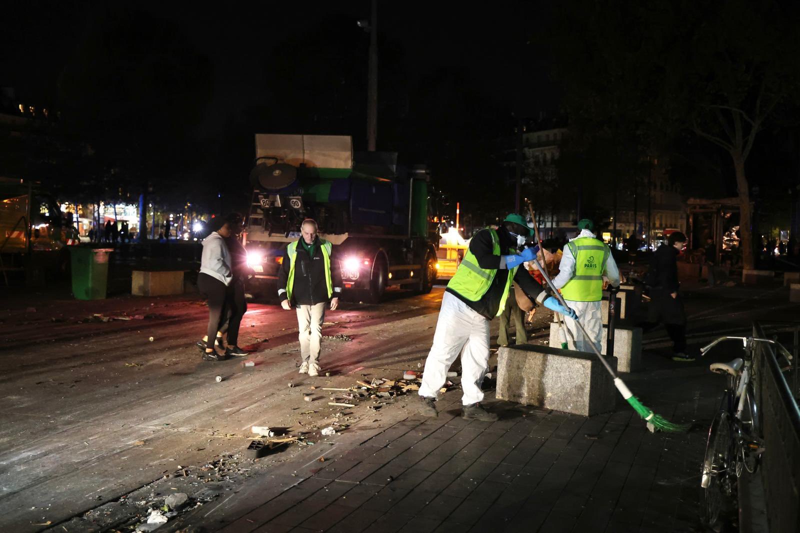 De nuit, des agents de propreté en gilet jaune balaient une place jonchée de débris, avec une benne derrière eux