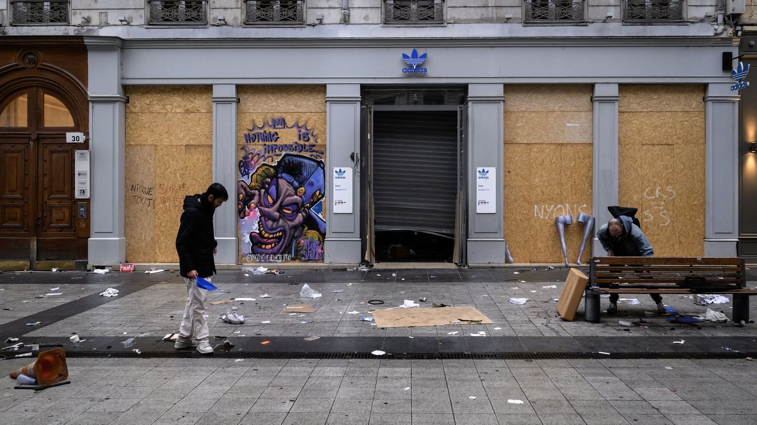 Un jeune homme ramasse des déchets devant une boutique aux vitrines condamnées par des planches, dans une rue de Lyon