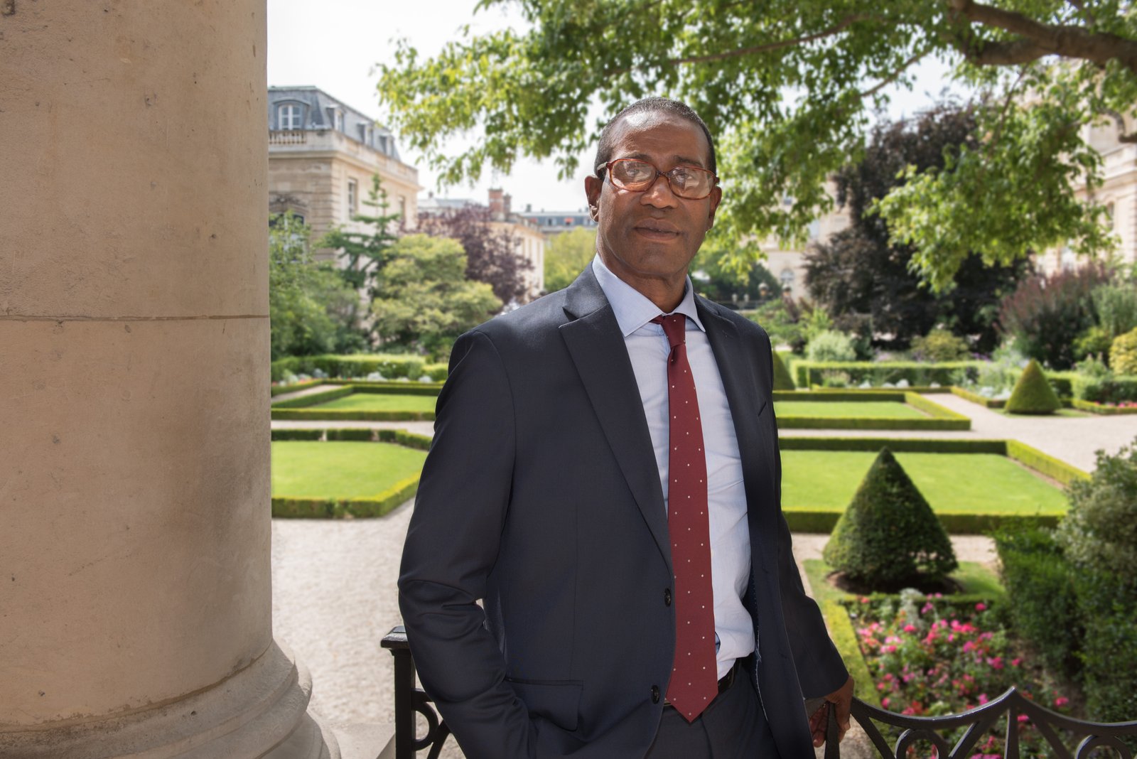 Max Mathiasin, lunettes et costume sombre, cravate bordeaux, pose souriant devant un jardin à la française