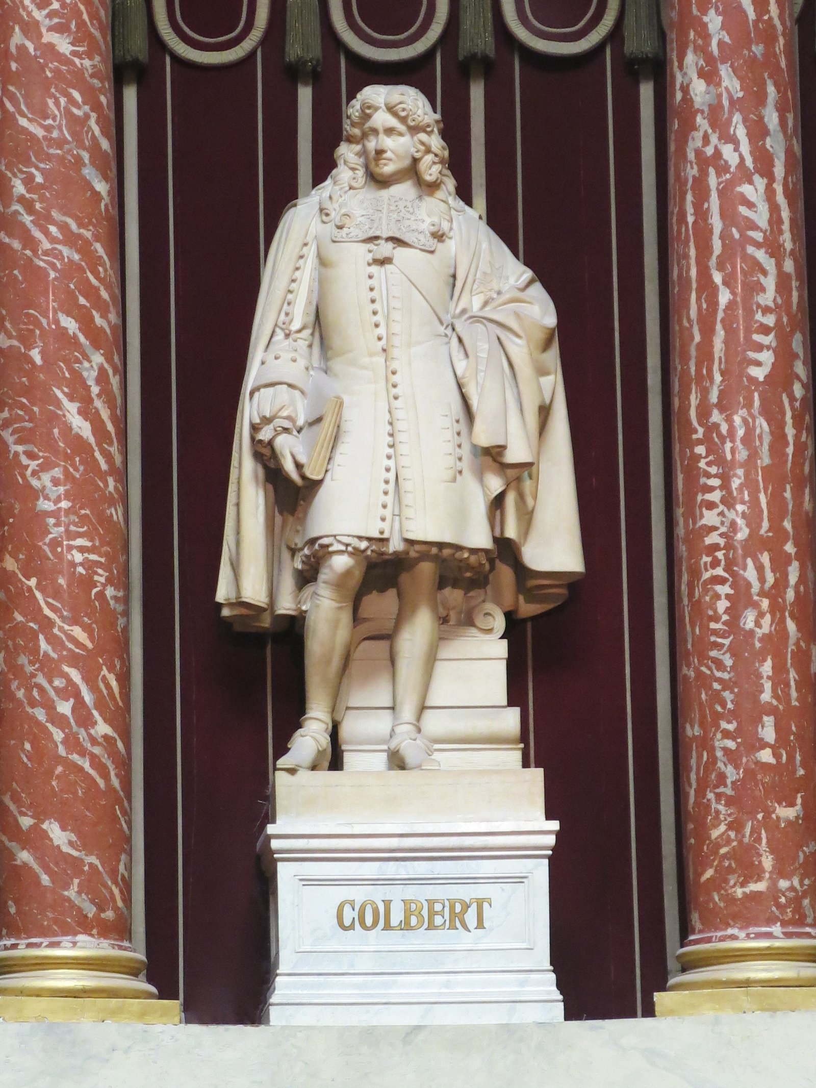 Statue de marbre blanc de Jean-Baptiste Colbert, en perruque et manteau, sur un socle gravé « Colbert », entre deux colonnes de marbre rouge