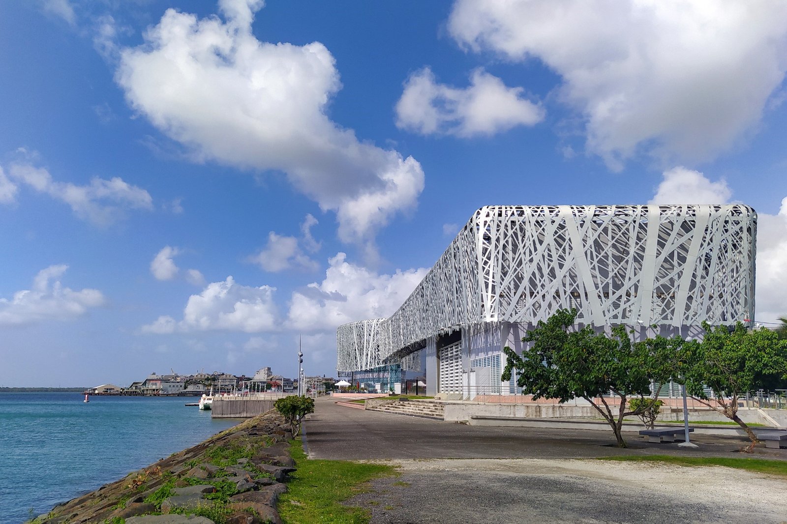 Le Mémorial ACTe de Pointe-à-Pitre, long bâtiment noir habillé d’une résille argentée, au bord de la mer sous un ciel bleu nuageux