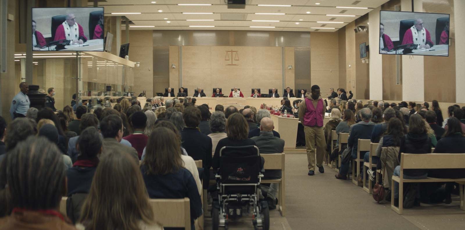 Une grande salle d’audience remplie de public, avec des écrans suspendus, dans une scène de la série