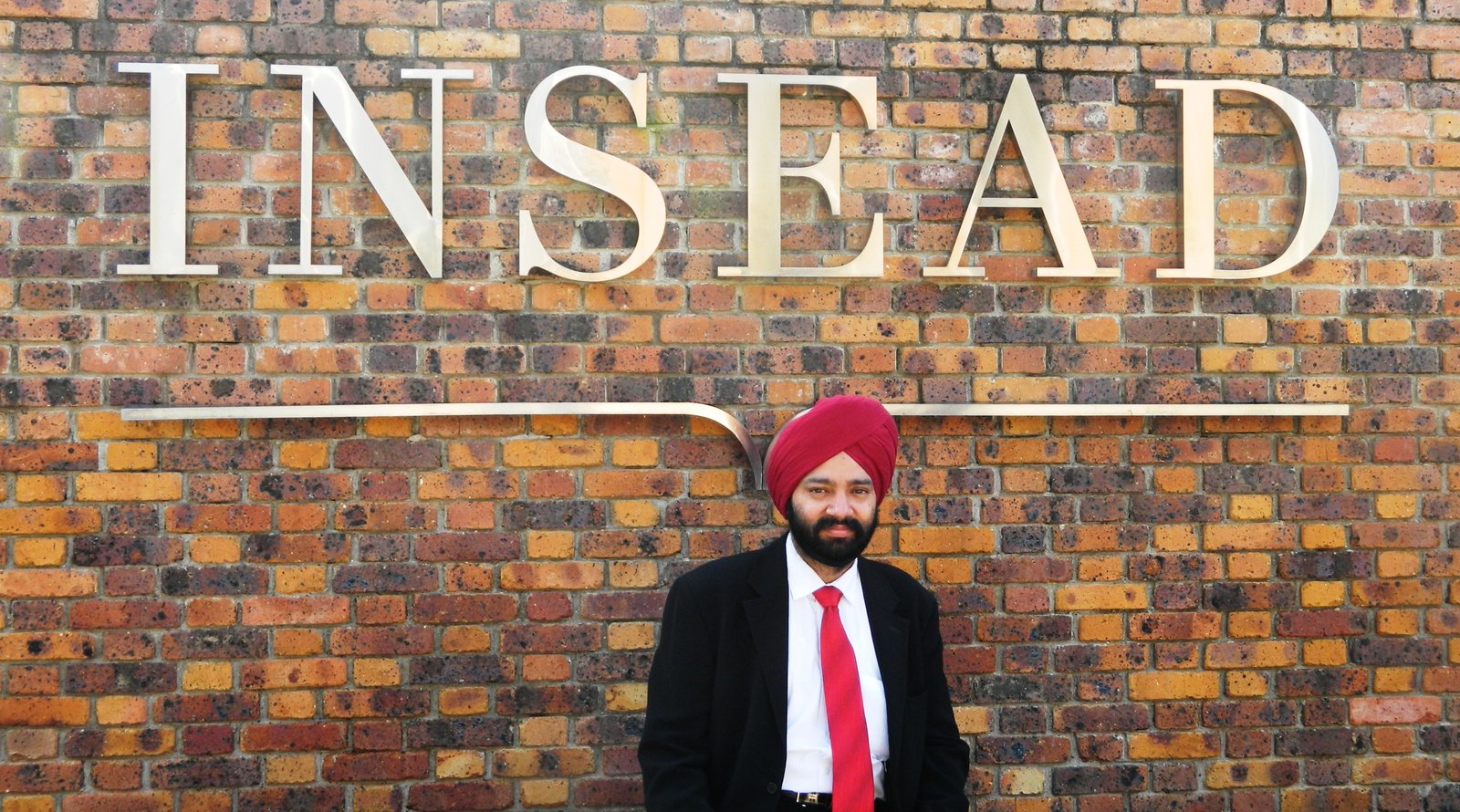 Un participant en costume et turban rouge pose devant le mur de briques marqué INSEAD