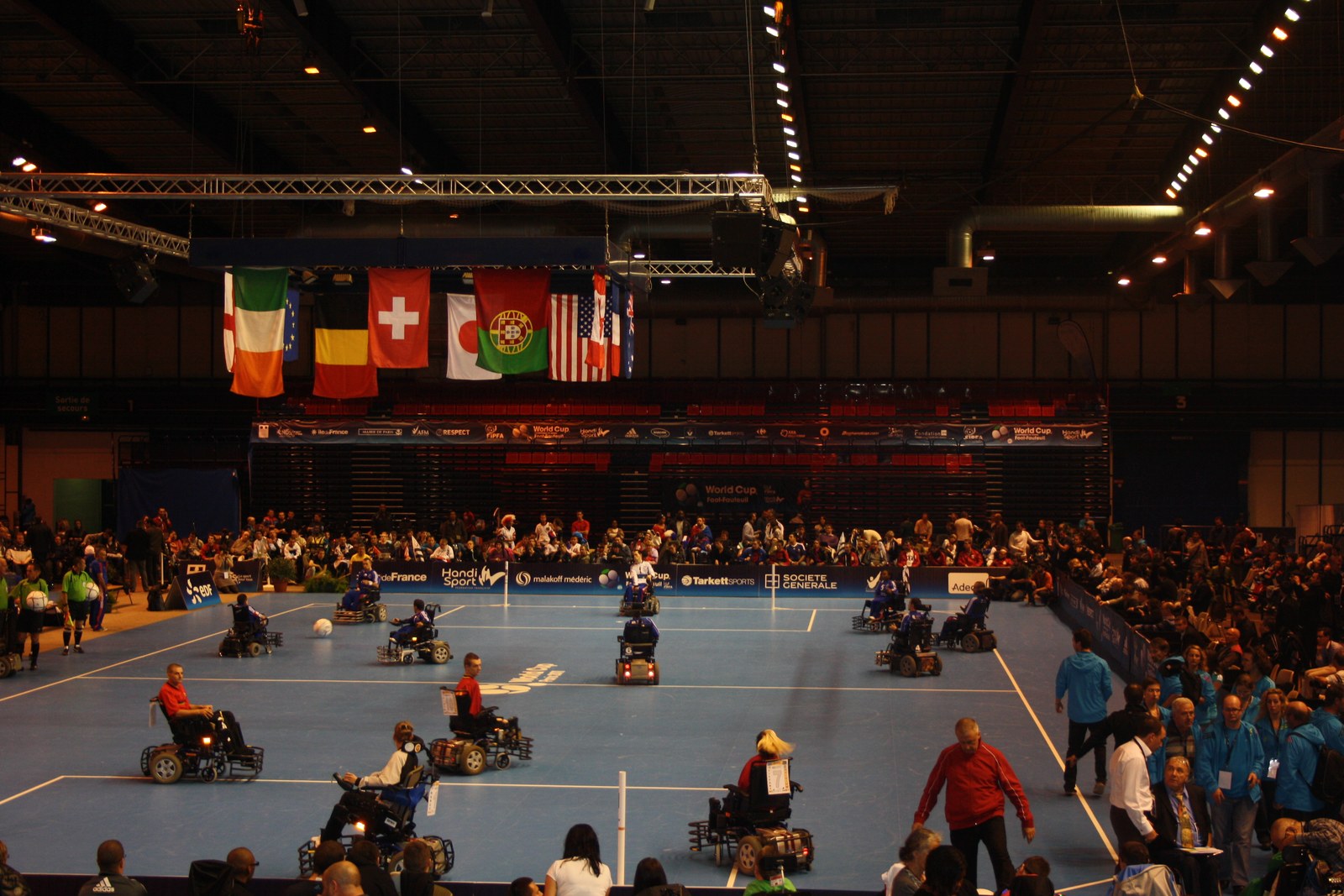 Vue d’ensemble d’un match de foot fauteuil dans une grande salle, sous les drapeaux des nations, devant des tribunes pleines