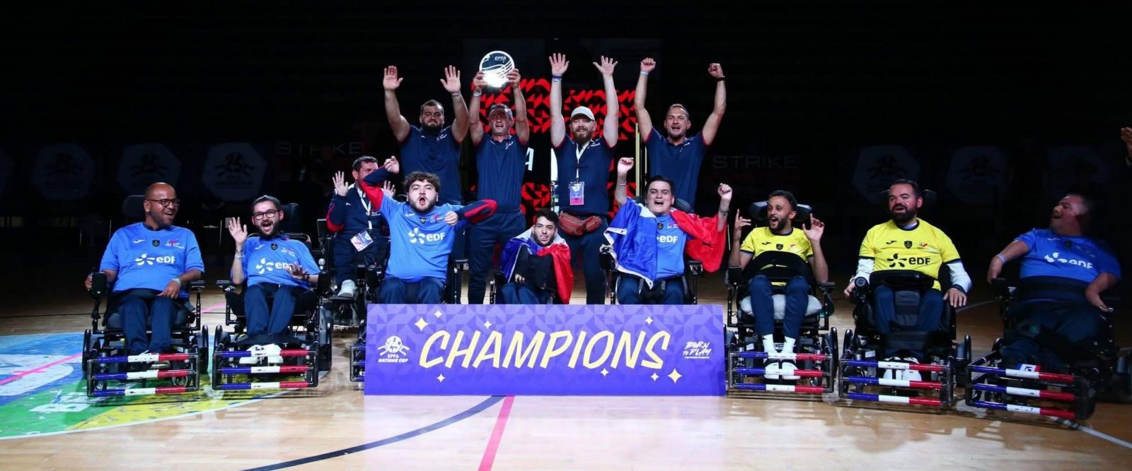 Les joueurs de l’équipe de France de foot fauteuil, en maillots bleus et jaunes, lèvent les bras derrière une banderole « Champions »