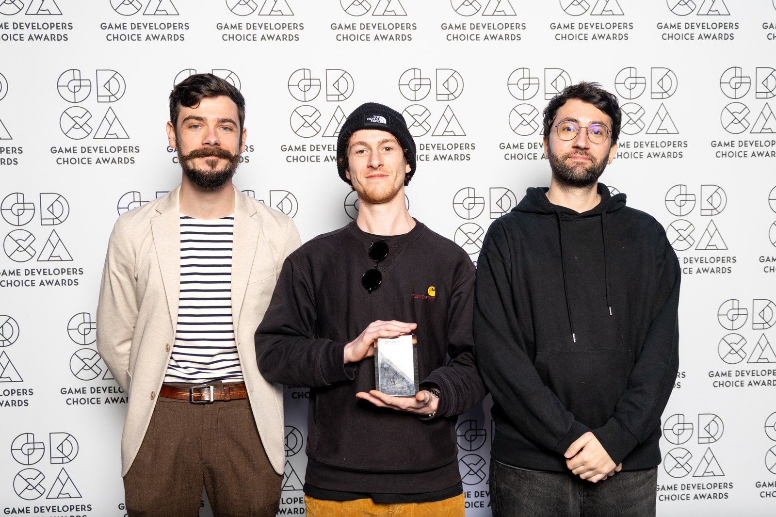 Trois membres de Sandfall Interactive, souriants, tiennent leur trophée du meilleur son devant le mur des Game Developers Choice Awards