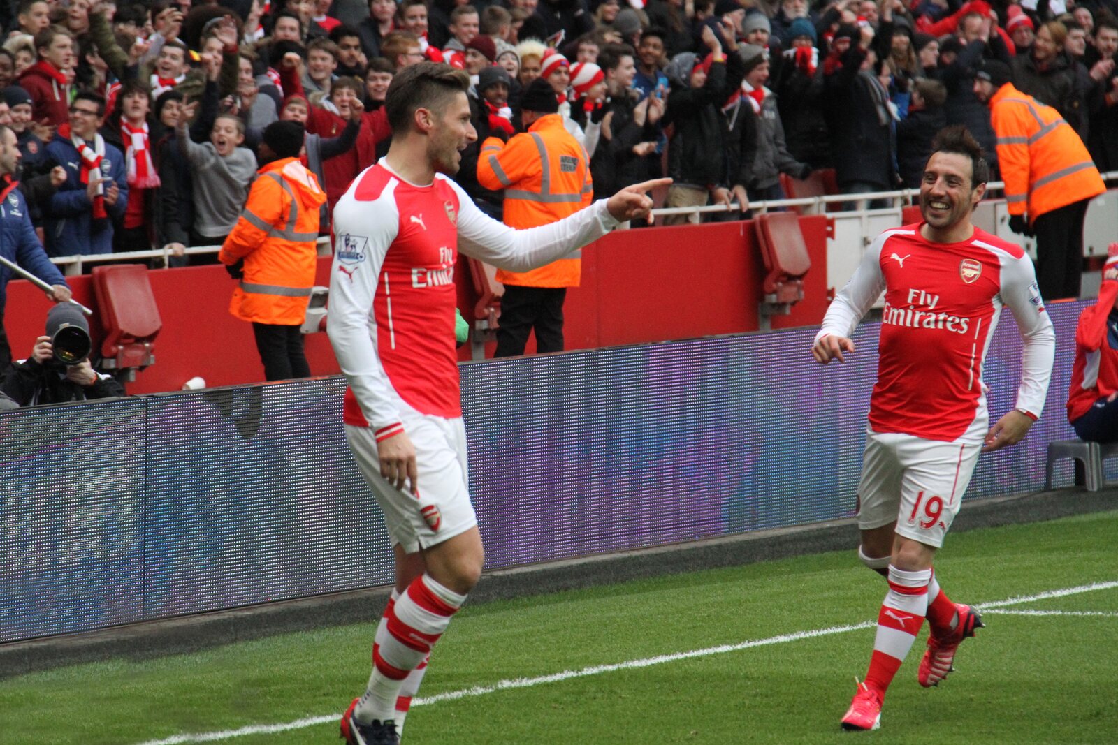 Olivier Giroud, maillot rouge et blanc d’Arsenal, pointe du doigt son coéquipier Santi Cazorla qui court vers lui, tribunes pleines derrière