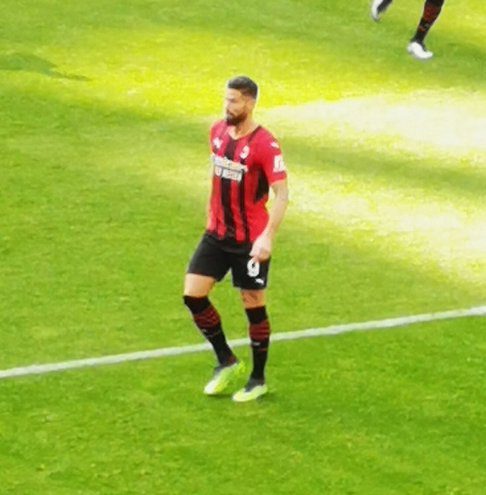 Olivier Giroud en maillot rouge et noir de l’AC Milan, debout sur la pelouse ensoleillée de San Siro