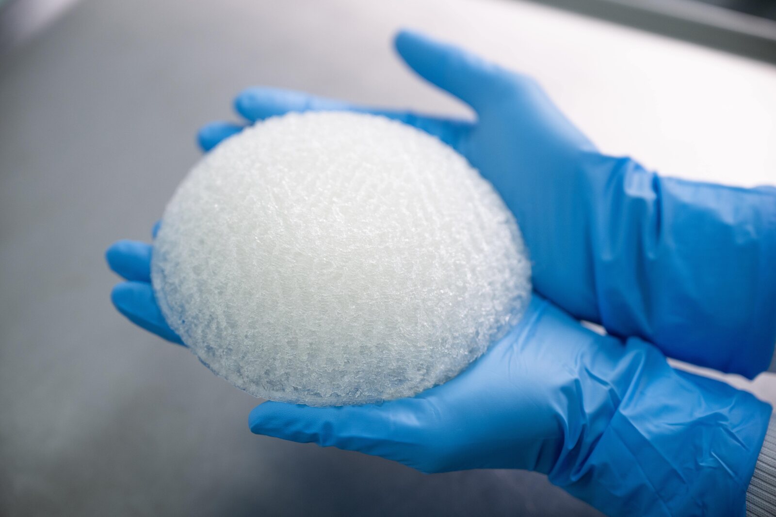 Deux mains gantées de bleu tiennent un dôme blanc et alvéolé d’hydrogel, de la taille d’un sein