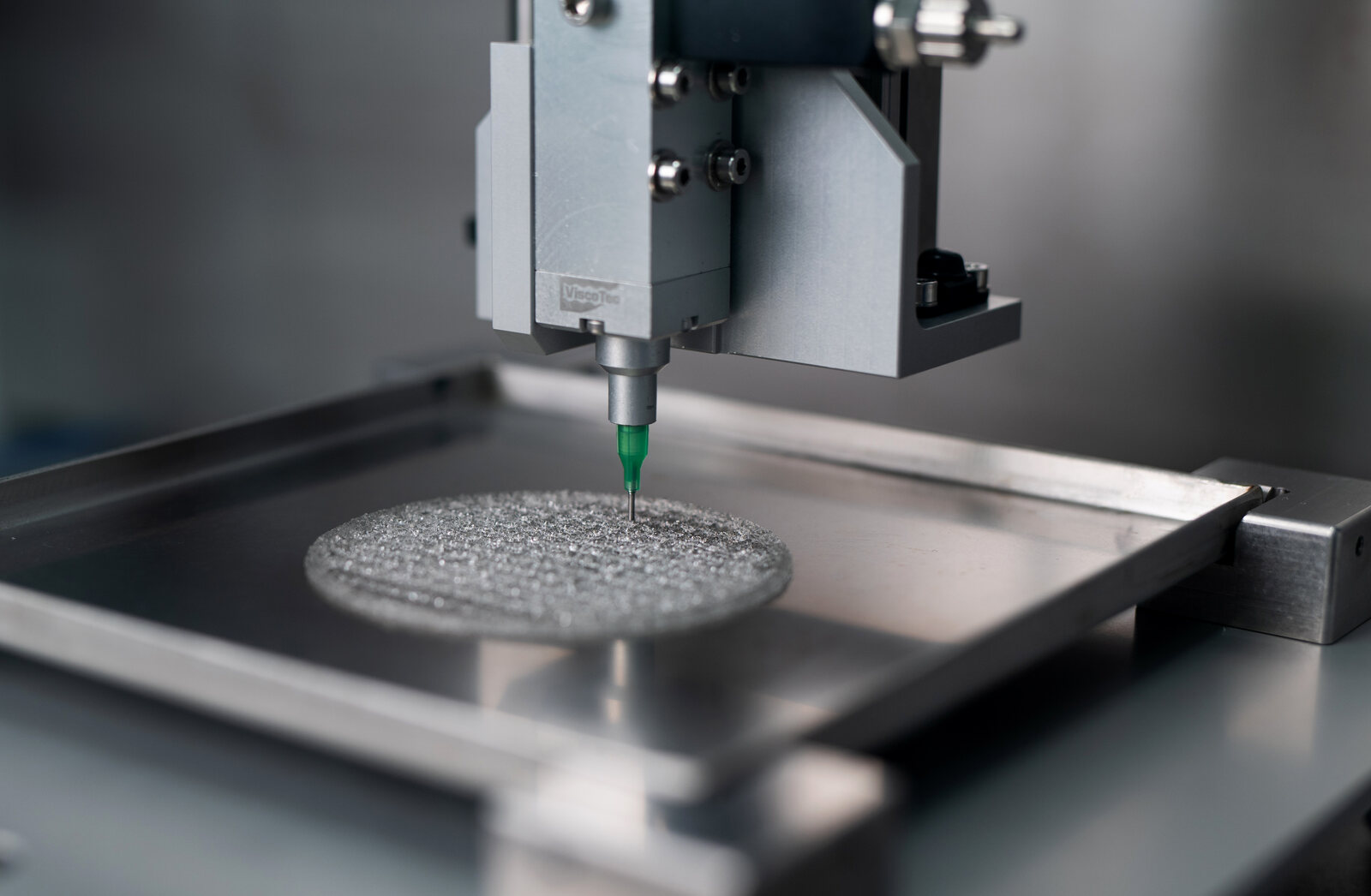 Une aiguille verte dépose de l’hydrogel sur un plateau métallique, formant une fine grille circulaire