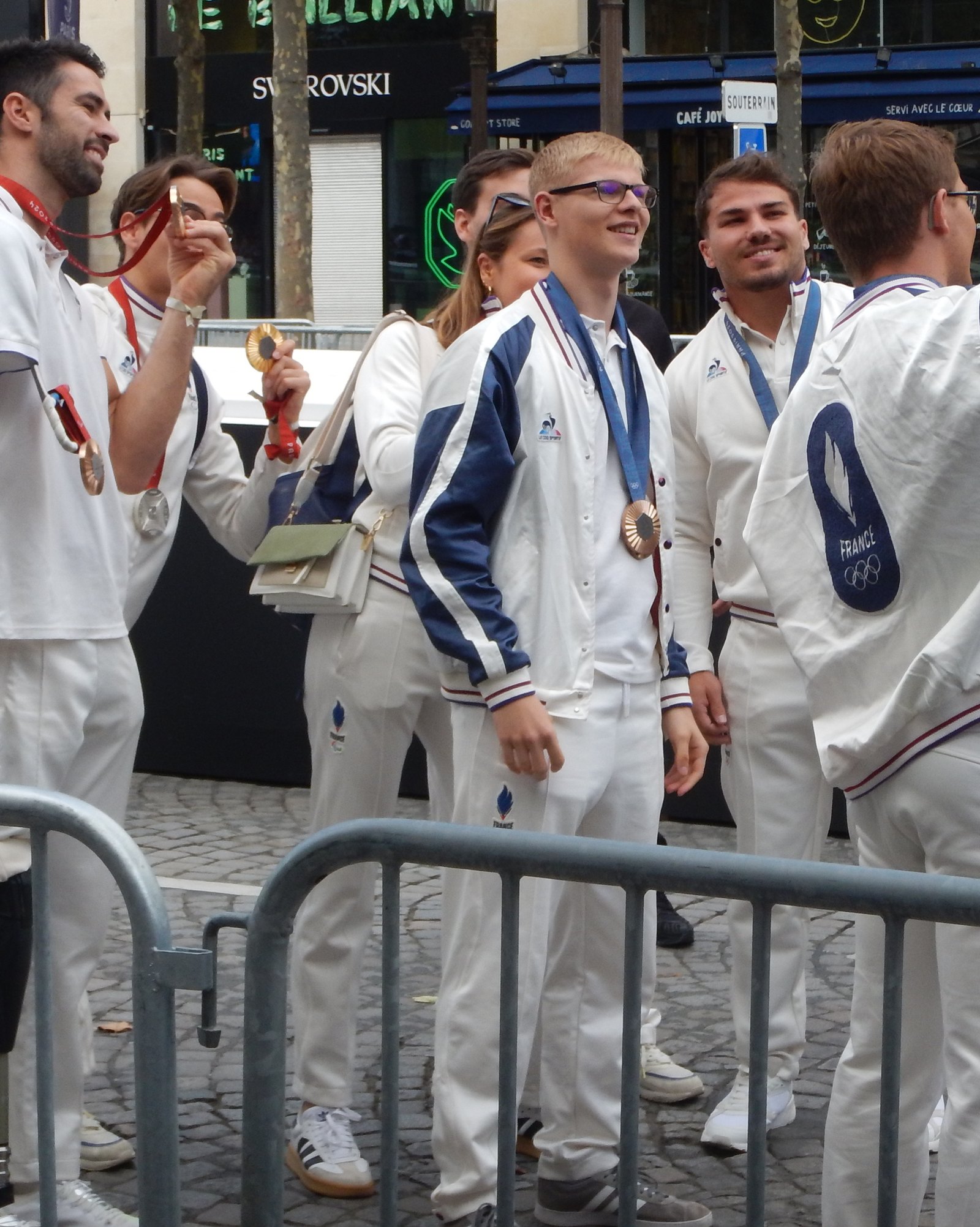Félix Lebrun souriant en veste blanche de l’équipe de France, médaille de bronze olympique autour du cou, entouré d’autres athlètes
