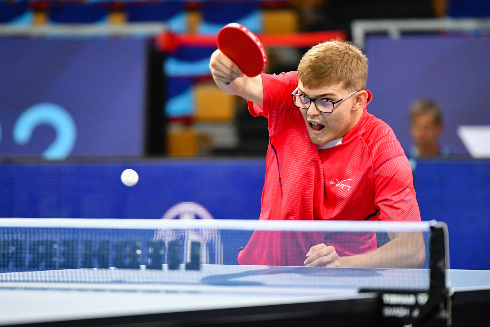 Alexis Lebrun en maillot rouge FFTT, lunettes, raquette prête, regard concentré au-dessus de la table