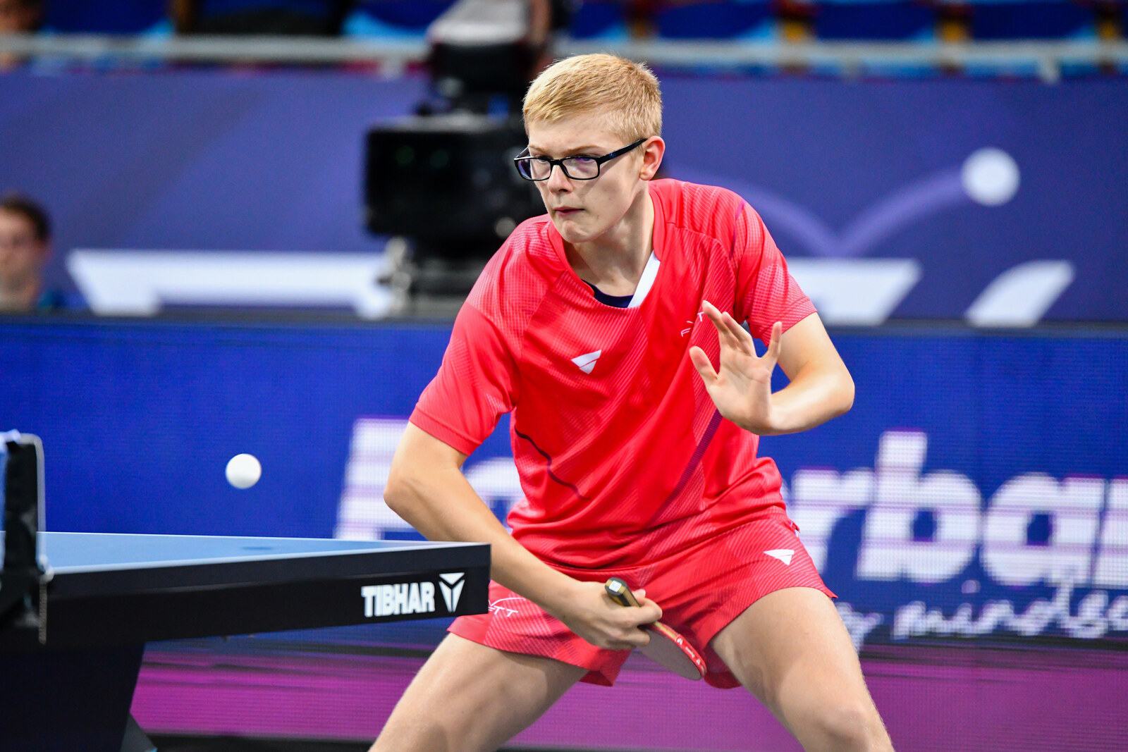 Félix Lebrun en maillot rouge, lunettes, raquette basse, balle en l’air au-dessus de la table bleue