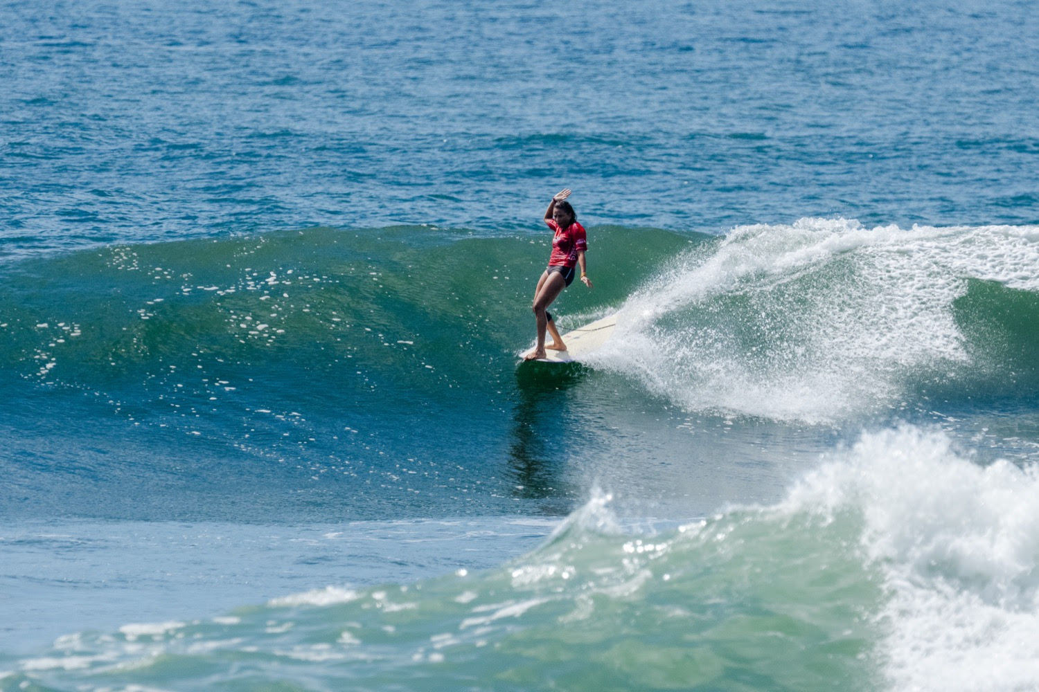 Alice Lemoigne, en lycra rouge, debout sur son longboard, glisse sur le haut d’une vague turquoise à El Sunzal
