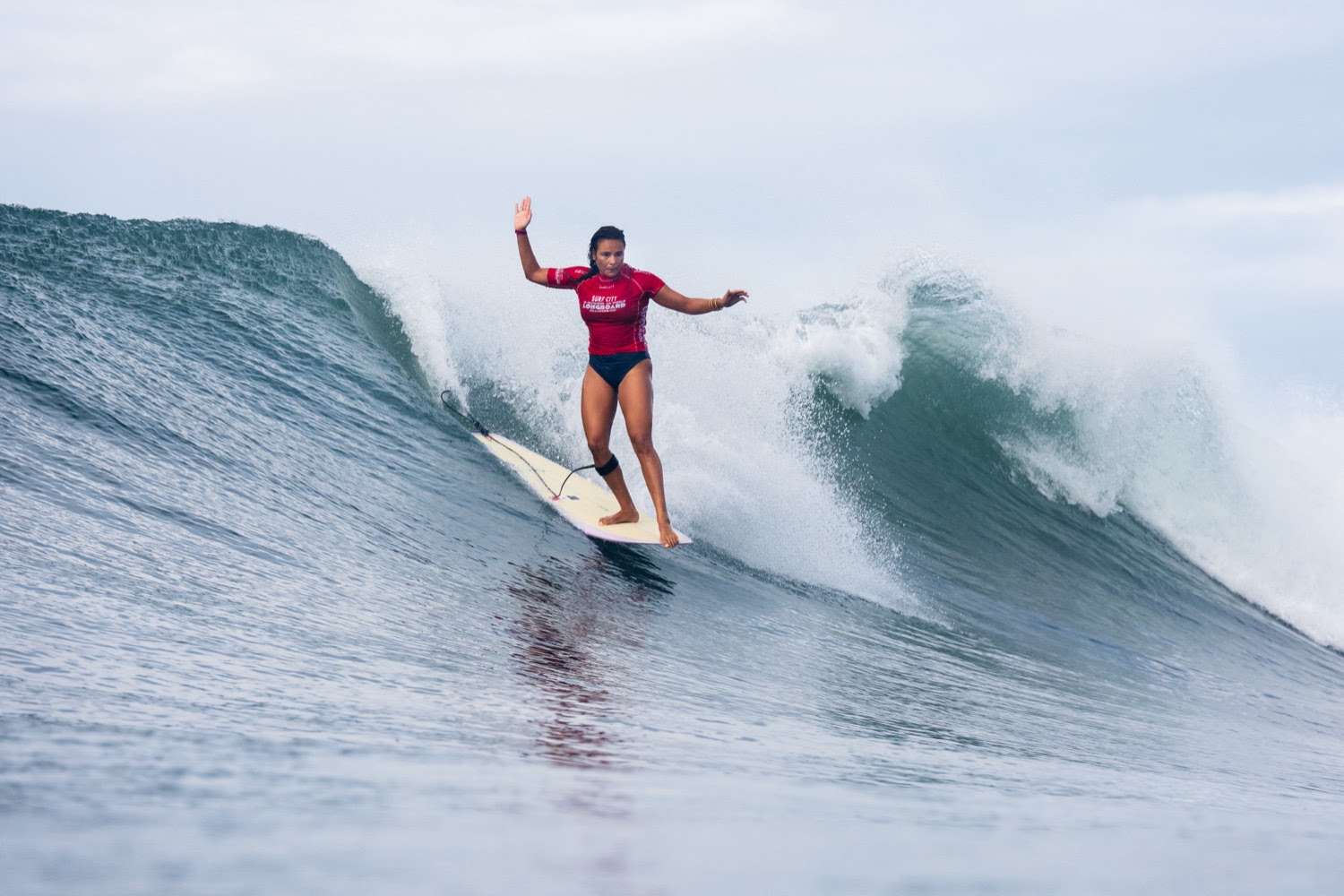Alice Lemoigne, bras écartés, file debout sur son longboard le long d’une vague qui se creuse derrière elle