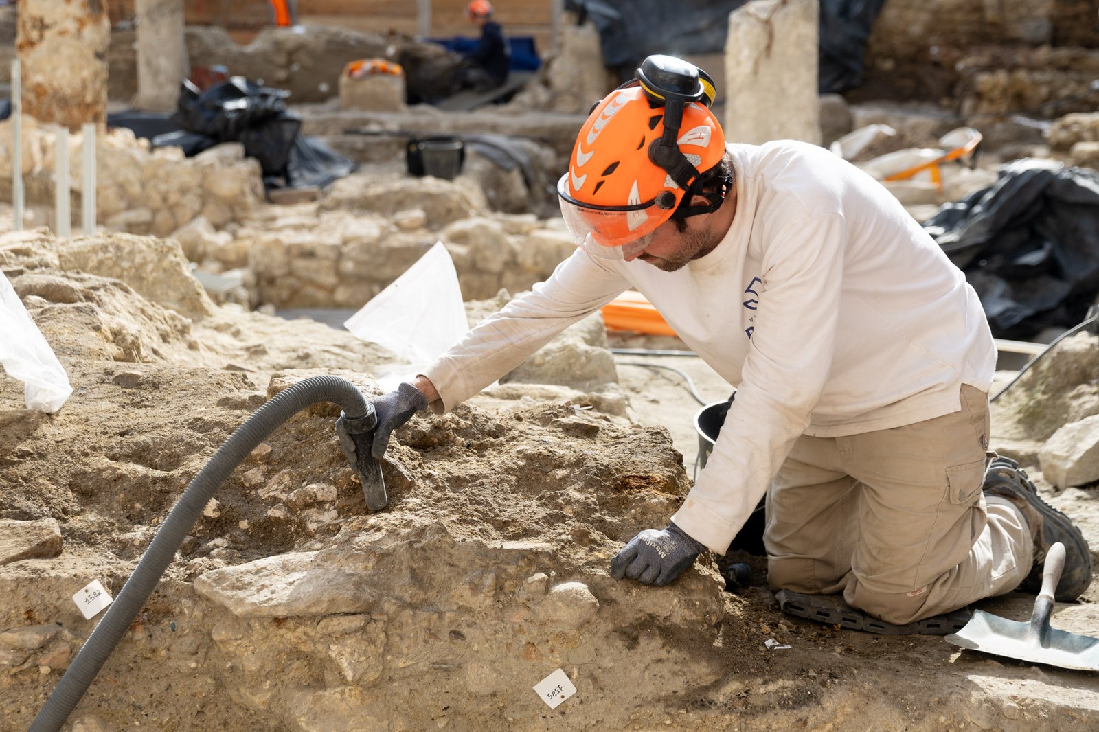 Un archéologue à genoux, casque orange et tee-shirt blanc, dégage la terre d’un vestige avec un aspirateur de chantier