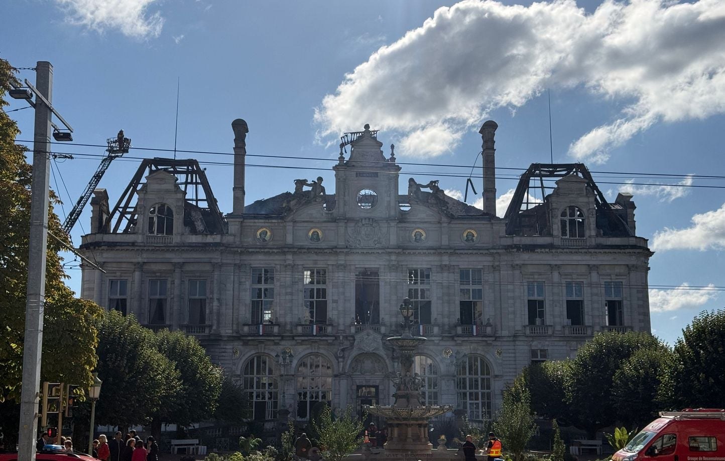 La façade de l’hôtel de ville de Limoges sous un ciel bleu, toiture effondrée, avec une grande échelle de pompiers et quelques passants devant la fontaine
