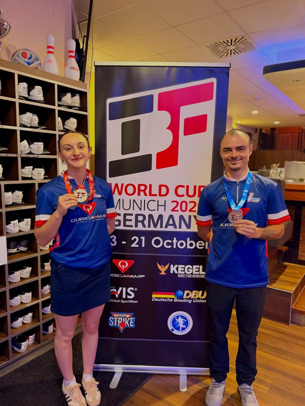 Manon Clément, médaille d’or, et Grégory Rius, médaille d’argent, sourient devant le panneau de la Coupe du monde IBF de Munich