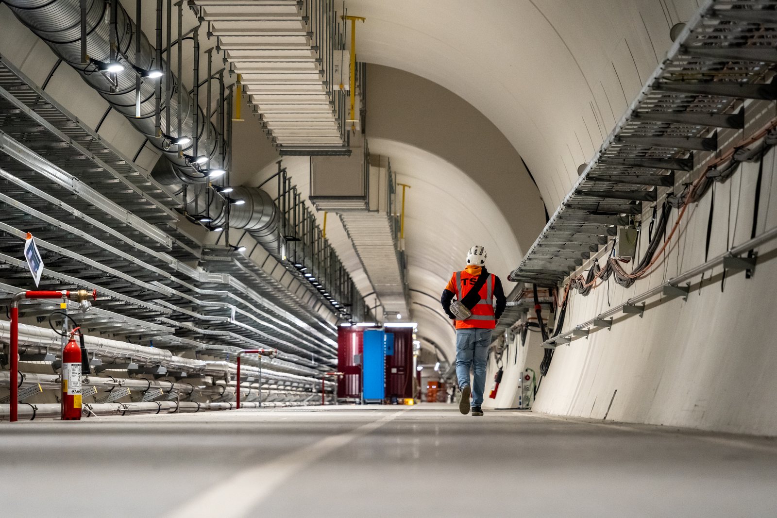 Un technicien en tenue orange marche seul dans un long tunnel blanc et neuf, bordé de chemins de câbles