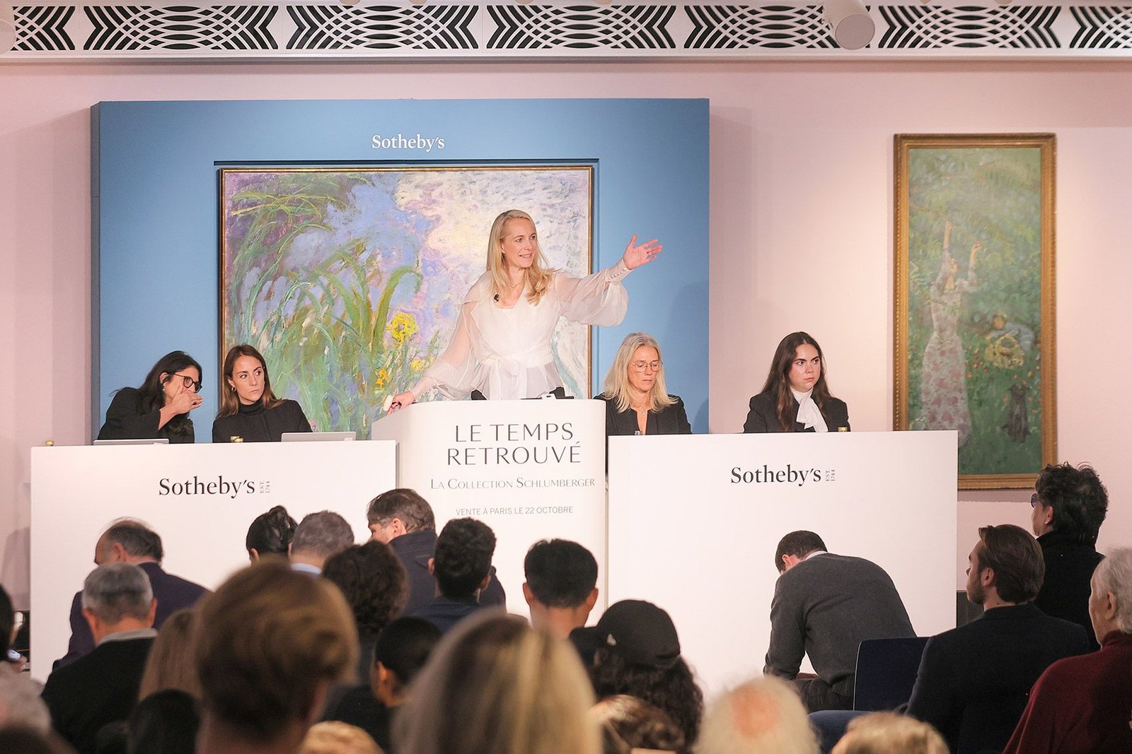Une commissaire-priseuse en robe claire, bras tendu, mène les enchères depuis un pupitre Sotheby’s, face à une salle comble, avec des téléphonistes à ses côtés