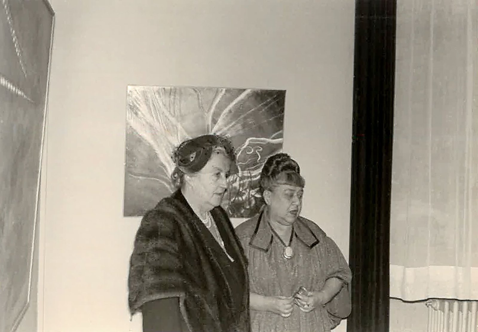 Photographie d’archives en noir et blanc : deux femmes âgées, en manteau, posent côte à côte devant une toile accrochée au mur