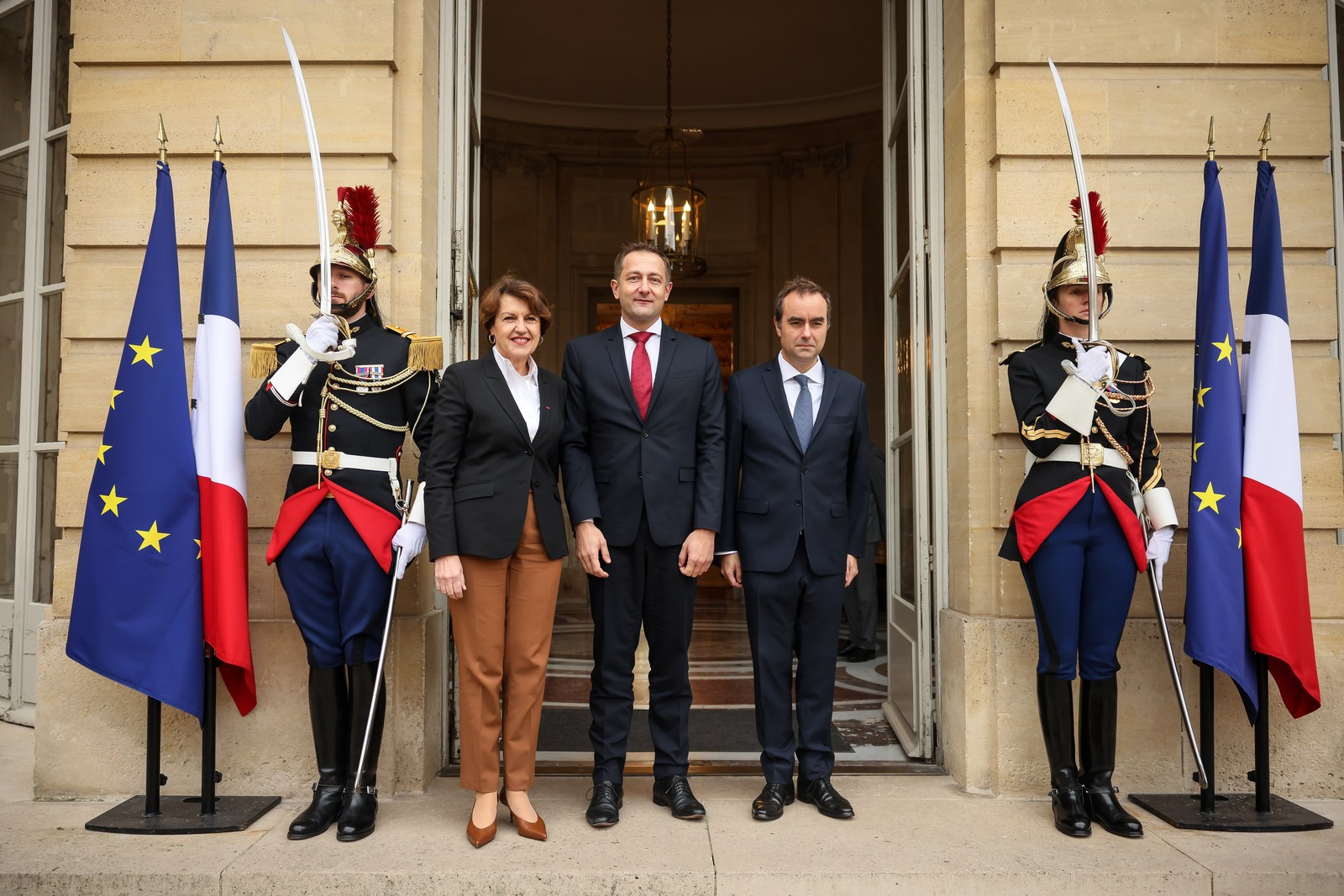 Sébastien Lecornu, Christophe Hansen et Annie Genevard posent sur un perron entre deux gardes républicains en grand uniforme et des drapeaux français et européens
