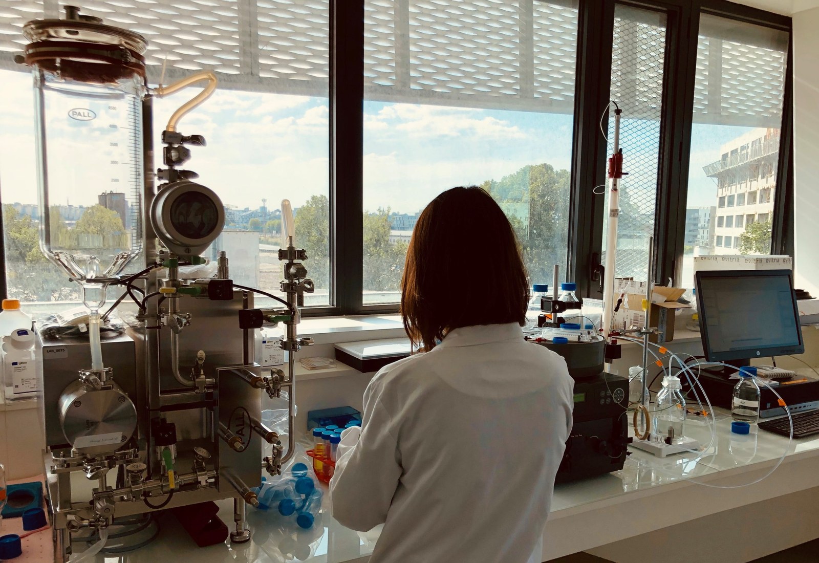 Une chercheuse de dos travaille devant un bioréacteur en inox, dans un laboratoire baigné de lumière