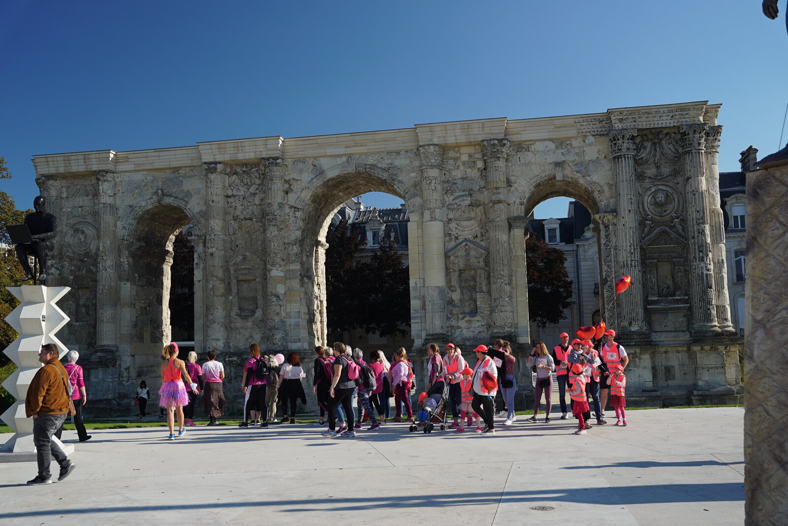 Des centaines de participantes en tee-shirt rose défilent devant la porte de Mars, arc romain de Reims, sous un ciel bleu