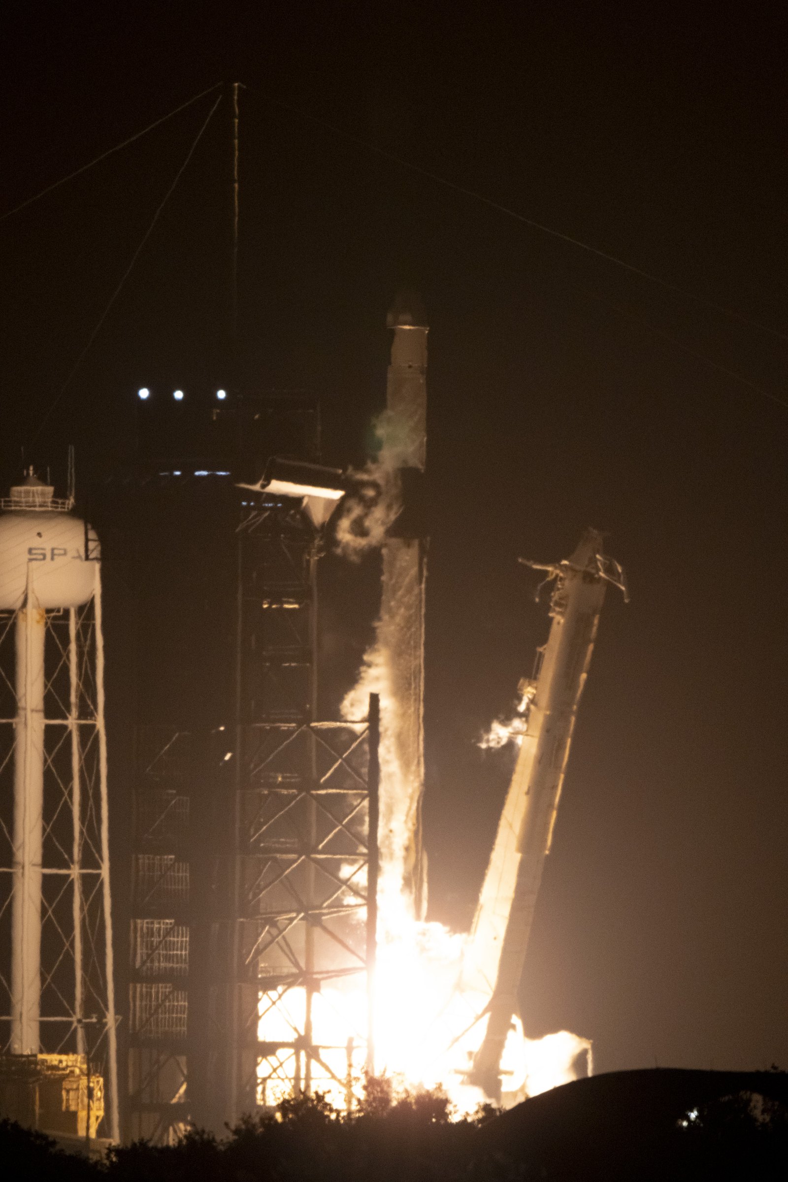 De nuit, une fusée Falcon 9 décolle dans un jet de flammes depuis son pas de tir, à côté de la tour de lancement