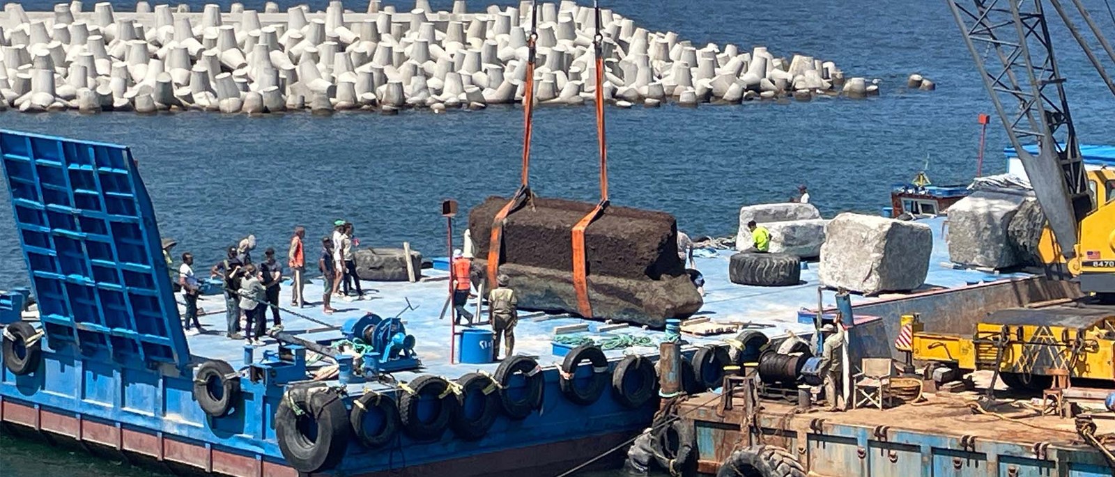 Un bloc de pierre du phare suspendu par des sangles orange au-dessus d’une barge bleue