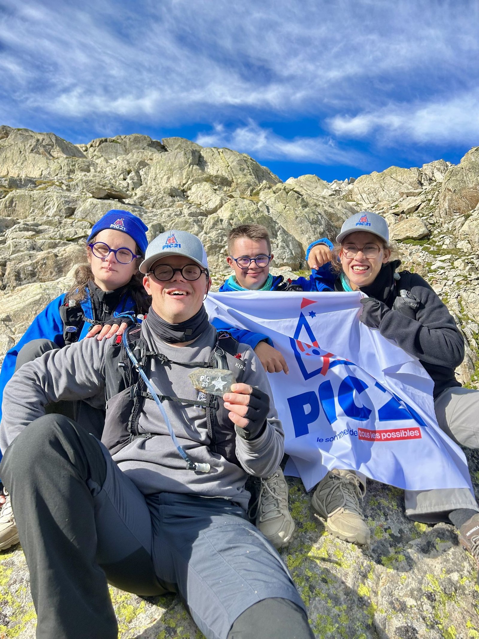 Quatre jeunes souriants, lunettes et casquettes, assis sur les rochers devant le drapeau Pic 21 ; au premier plan, l’un d’eux tient une pierre dans sa main