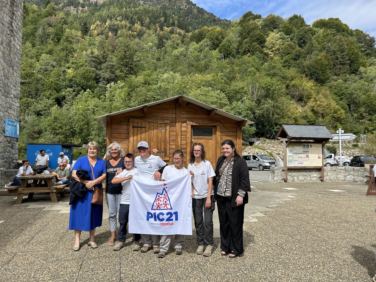 Une dizaine de personnes souriantes posent devant un chalet de bois en montagne, en tenant une banderole Pic 21