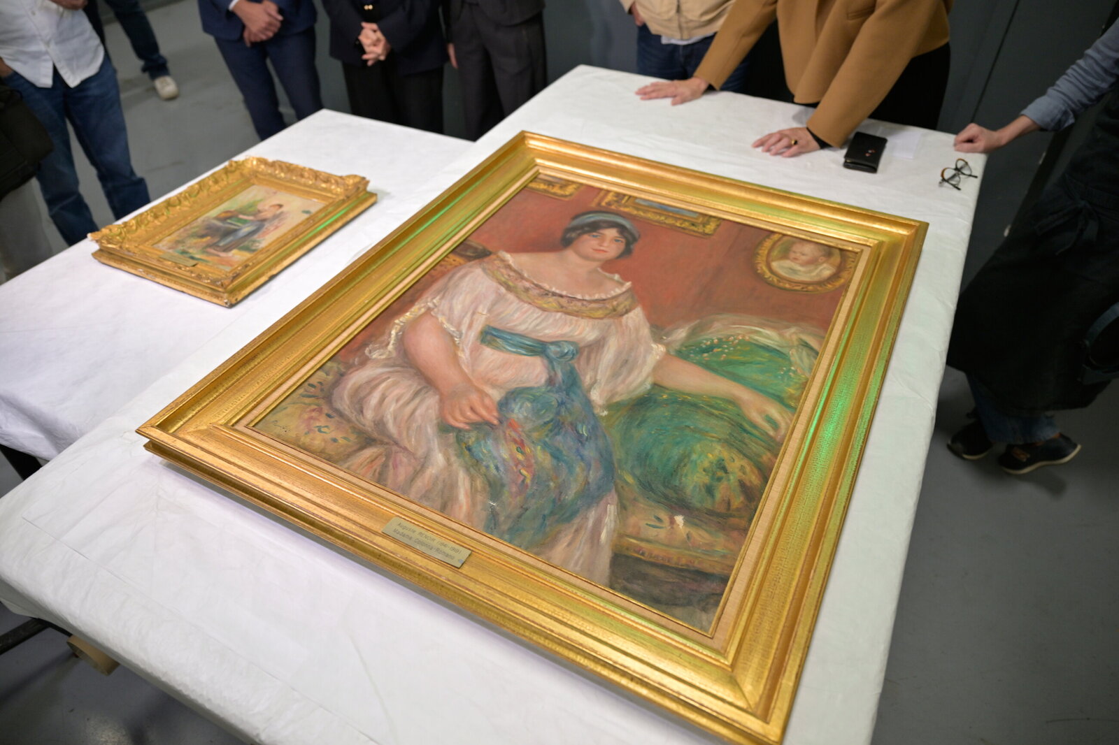 Des visiteurs entourent « Portrait de Madame Colonna Romano », grand tableau au cadre doré posé à plat sur une table blanche, avec le petit « Femme au puits » à l’arrière-plan