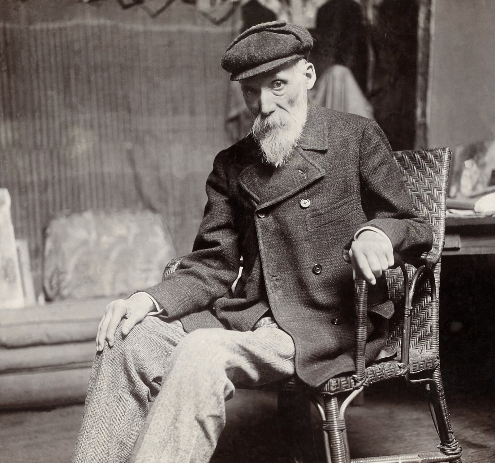 Photographie en noir et blanc de Pierre-Auguste Renoir âgé, barbe blanche et casquette, assis dans un fauteuil en osier
