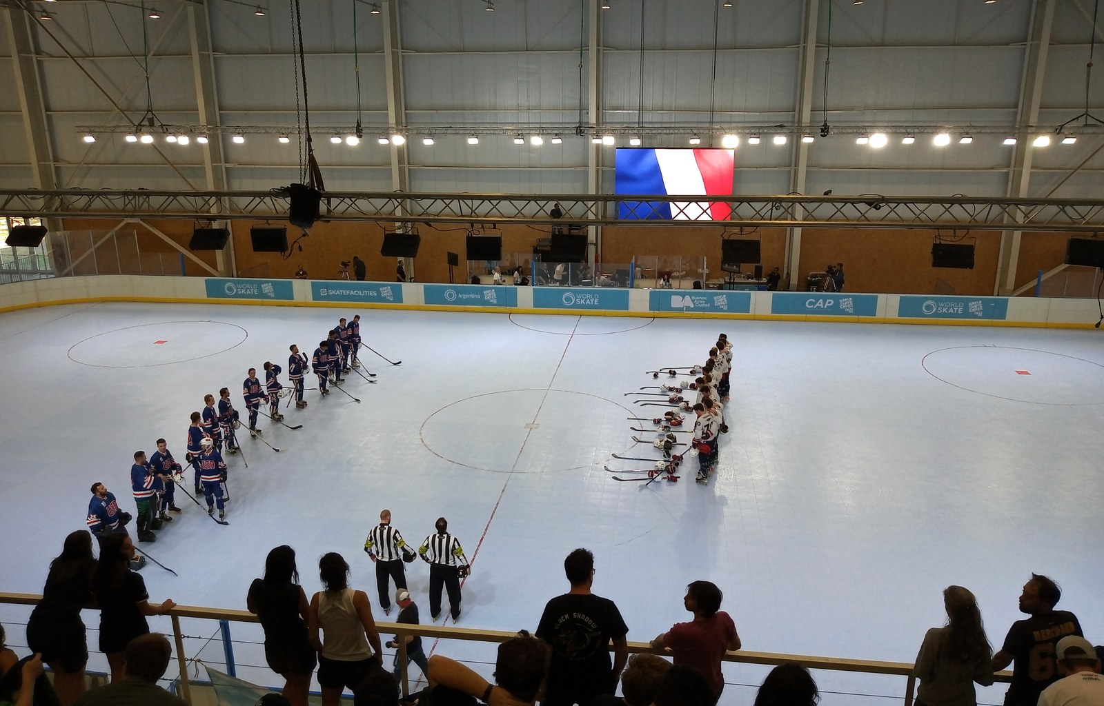 Deux équipes de roller hockey alignées sur une piste, drapeau français affiché sur un écran géant au fond