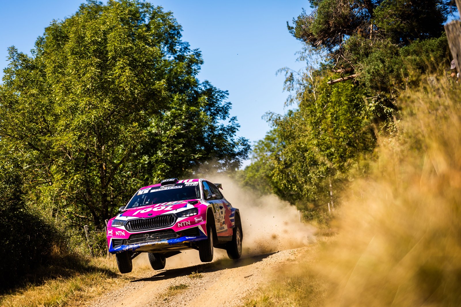 La Škoda Fabia rose et blanche de Sarah Rumeau soulève la poussière sur une piste en forêt, sous un ciel bleu
