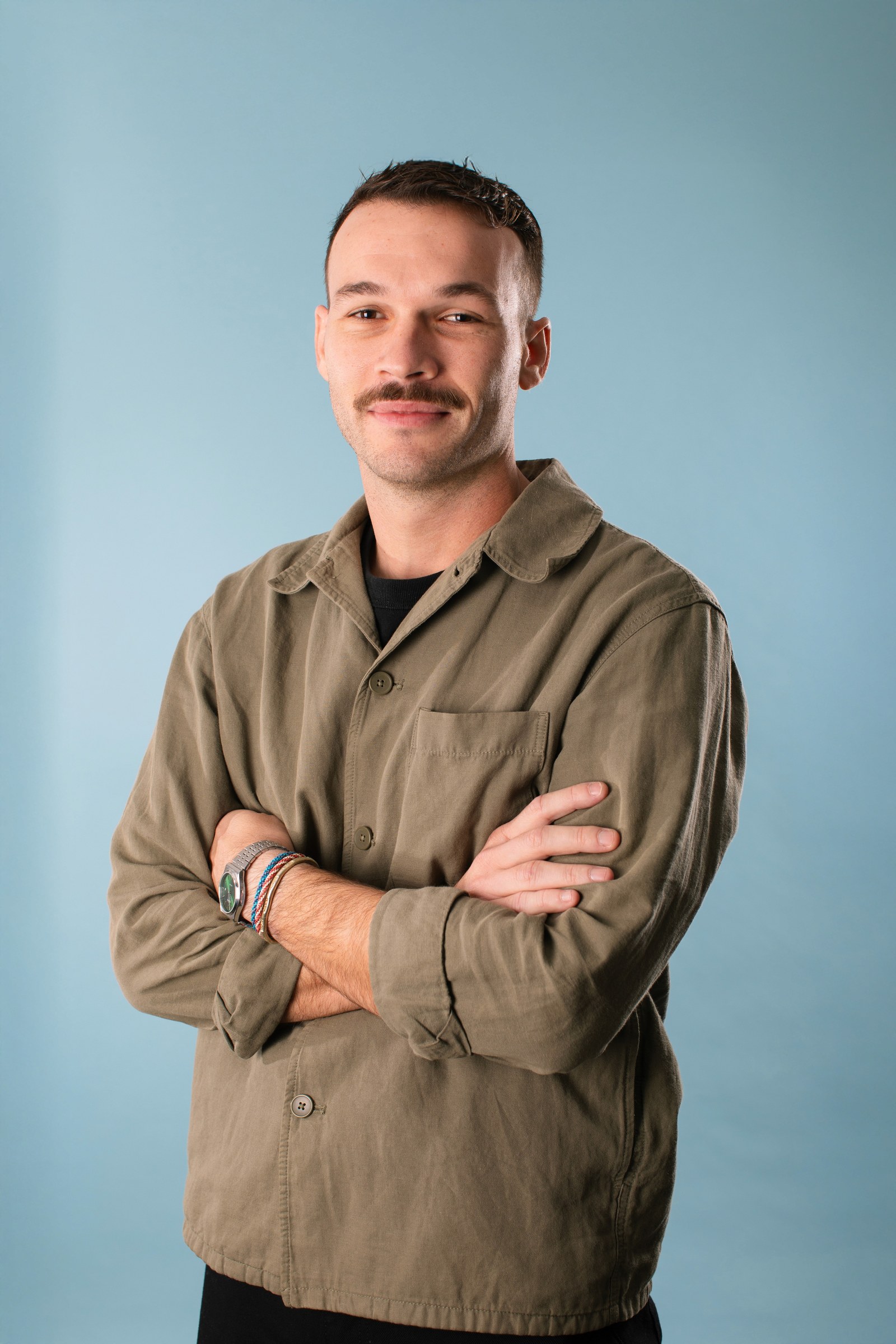 Portrait de presse de Dorian Barrère, bras croisés, chemise olive, fond bleu clair