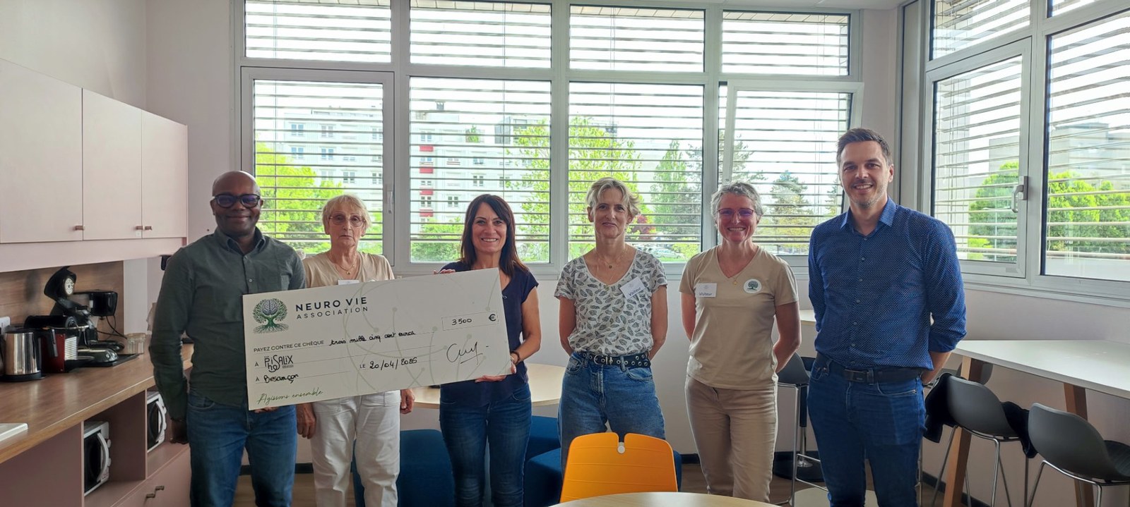 Six personnes souriantes dans une salle lumineuse, dont deux tiennent un grand chèque de l’association Neuro Vie