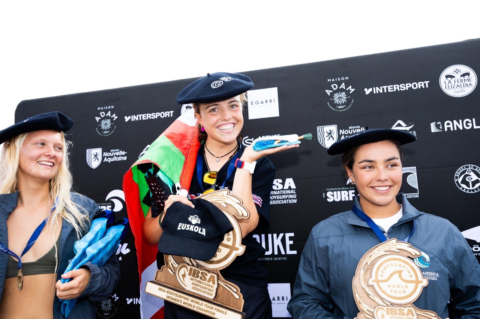 Uhaïna Kailani Hegoas, béret basque sur la tête et drapeau à la main, sourit au centre du podium, entourée de deux concurrentes