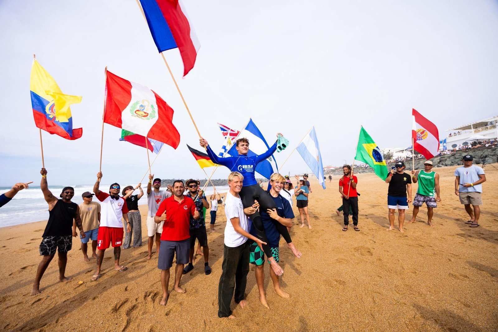 Niels Schorsch, en lycra bleu, porté en triomphe par deux jeunes au milieu des drapeaux de nombreux pays sur la plage