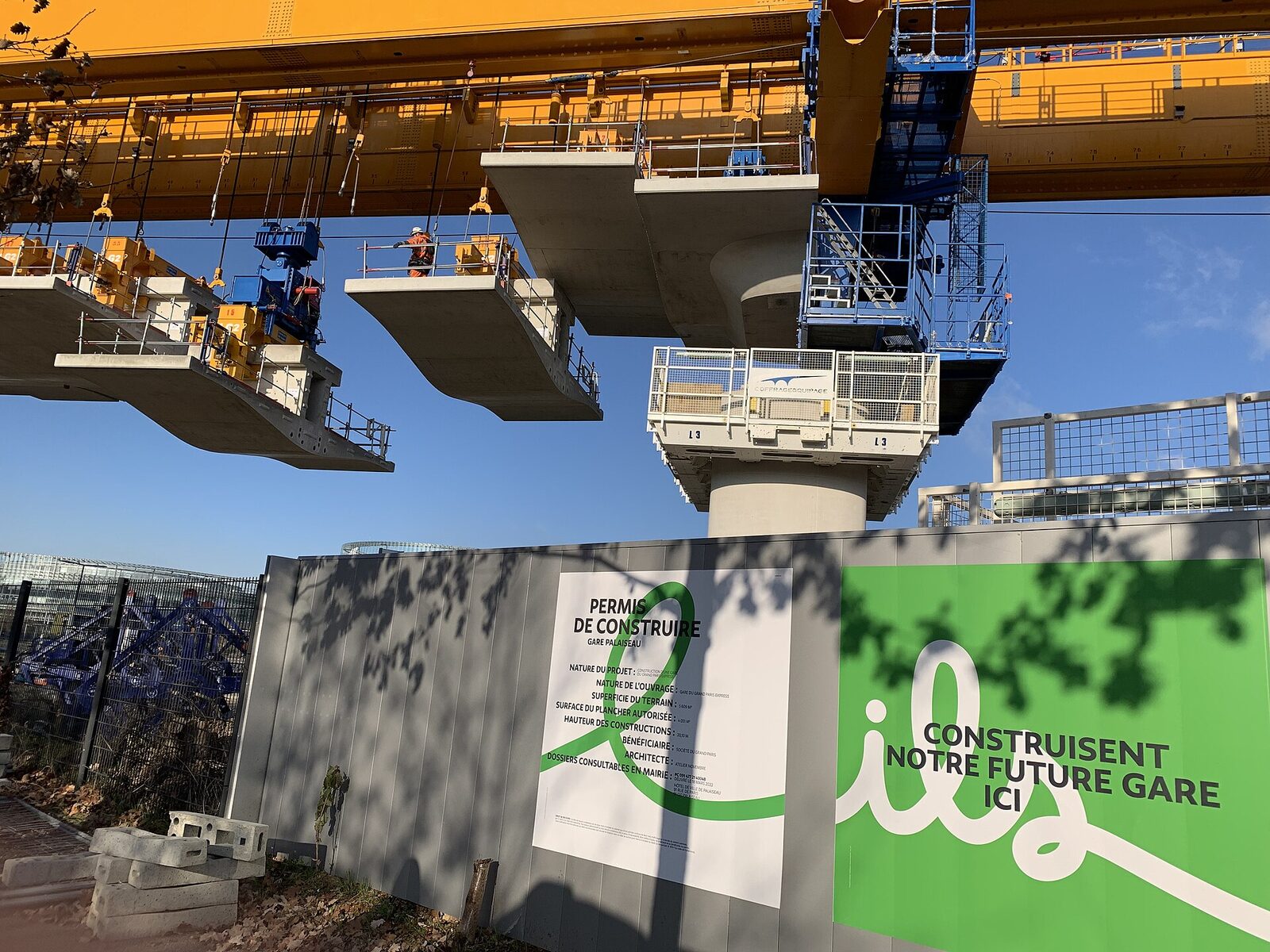 Un voussoir de béton suspendu sous une grande poutre de lancement jaune, au-dessus d’une palissade de chantier