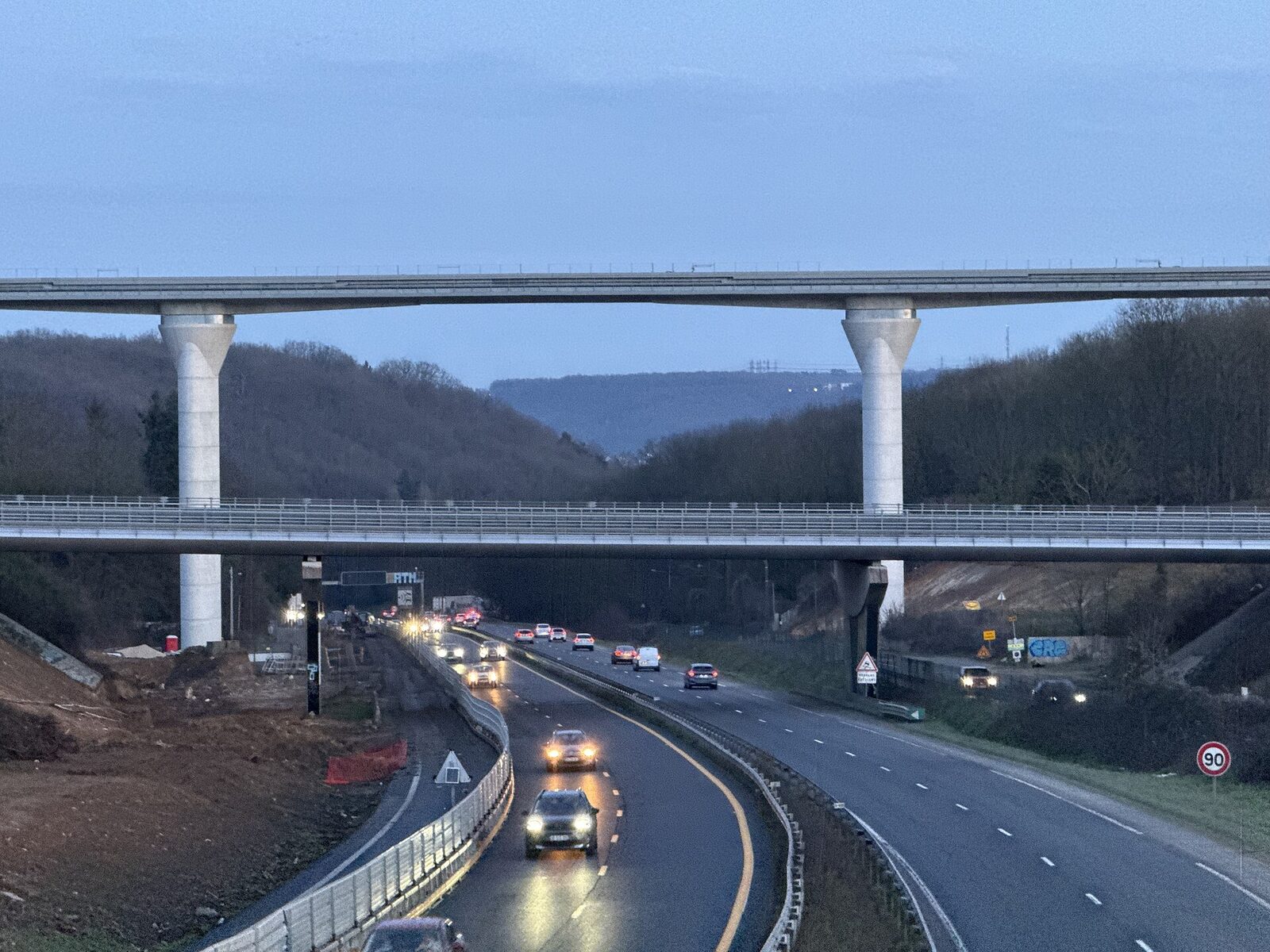 Le viaduc franchit une vallée boisée au-dessus d’une voie rapide où circulent des voitures phares allumés