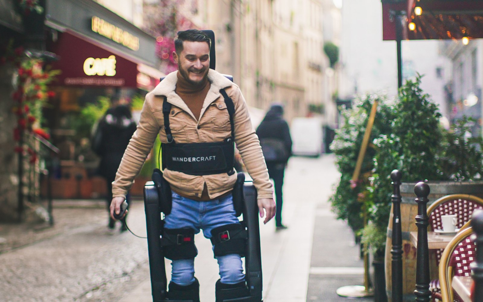 Un homme souriant marche dans une rue piétonne, sanglé dans un exosquelette Wandercraft, devant la terrasse d’un café