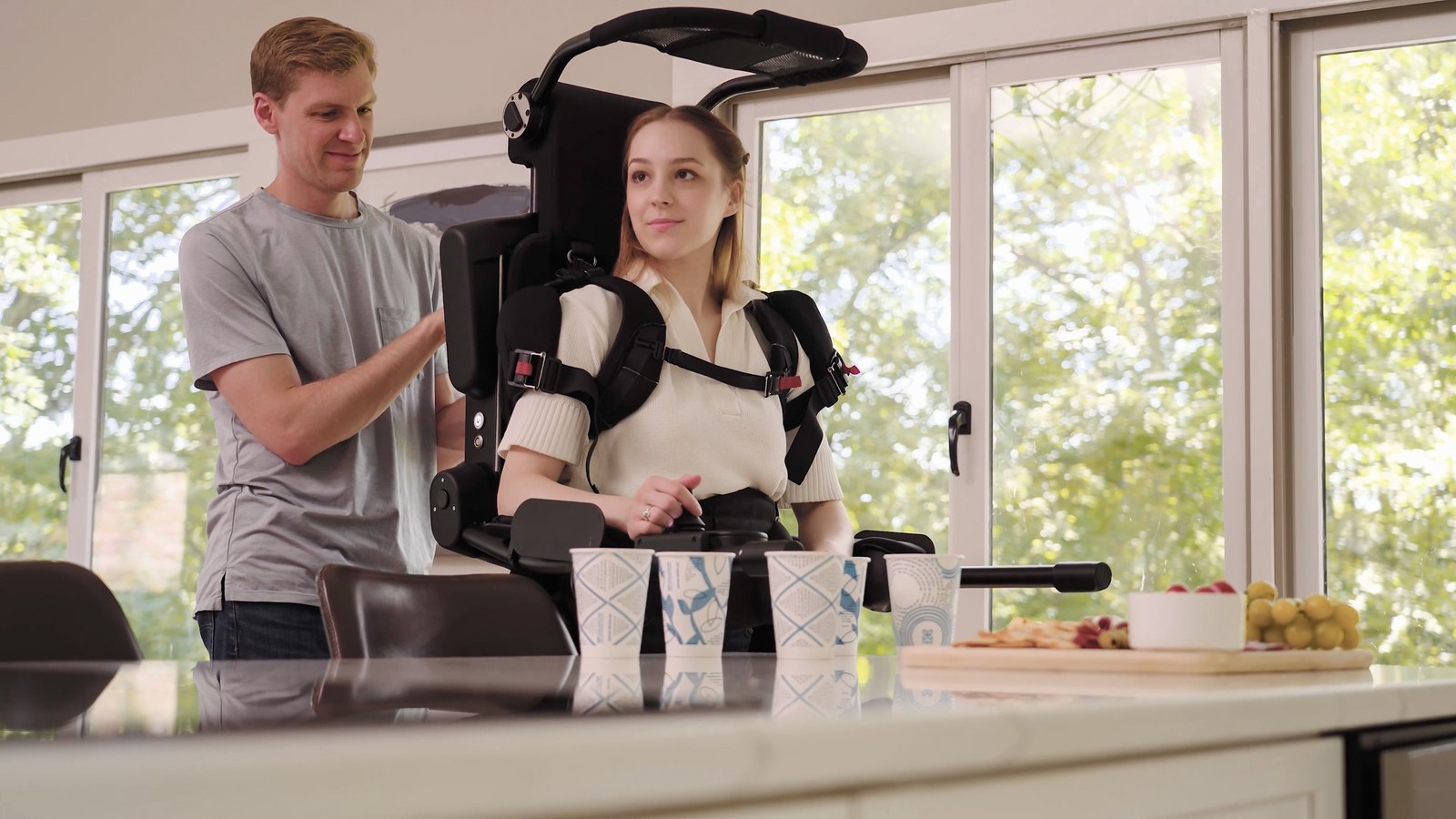 Une jeune femme debout dans l’exosquelette Eve, derrière un plan de travail de cuisine, aidée par un homme qui ajuste une sangle
